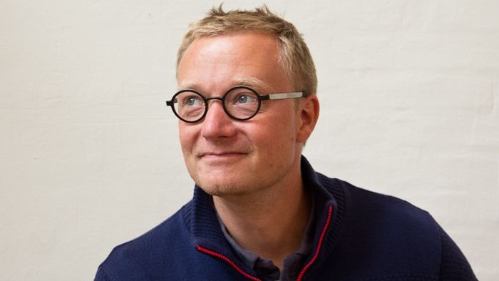 En smilende mand iført runde briller, en marineblå sweater med rød kant og en lys skjorte.