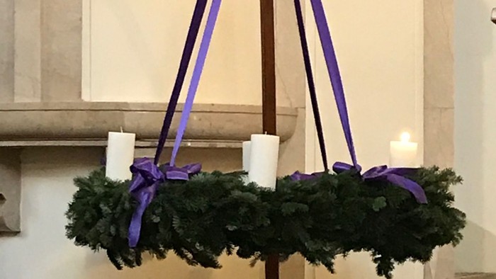 Ein Adventskranz mit drei brennenden Kerzen und lila Bändern vor einem steinernen Hintergrund