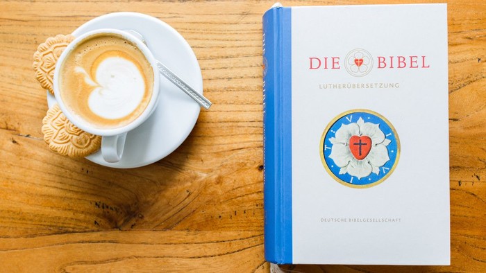 Eine Kaffeetasse mit Latte Art neben einer Luther-Bibelübersetzung auf einem Holztisch.