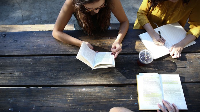 Zwei Frauen sitzen auf einem hölzernen Steg, jede vertieft in ein Buch. Die eine trägt ein gelbes Hemd, die andere hat lange Haare.