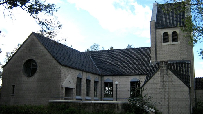 Ansicht der Kirche St. Kunigunde in Sennestadt mit hellem Ziegelmauerwerk, dunklem Satteldach und markantem Kirchturm. Die Kirche steht umgeben von Bäumen und Grünanlagen unter blau-weißem Himmel.