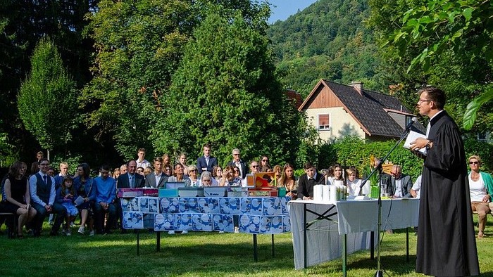 Ein Priester spricht vor einer Menschenmenge im Freien, im Hintergrund ein Haus und Bäume.