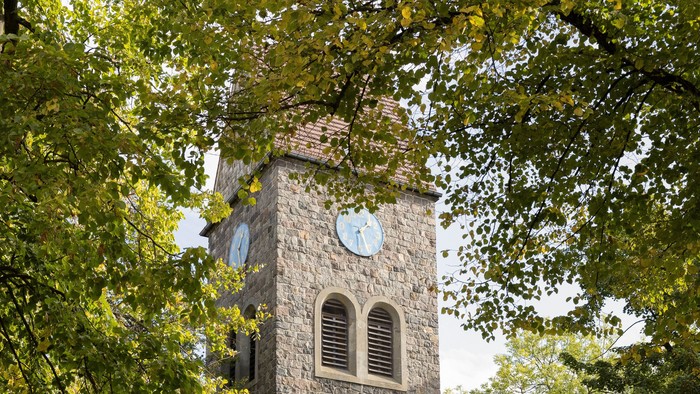 Der Kirchturm aus Stein mit Uhr und Fenster steht in einem Park.
