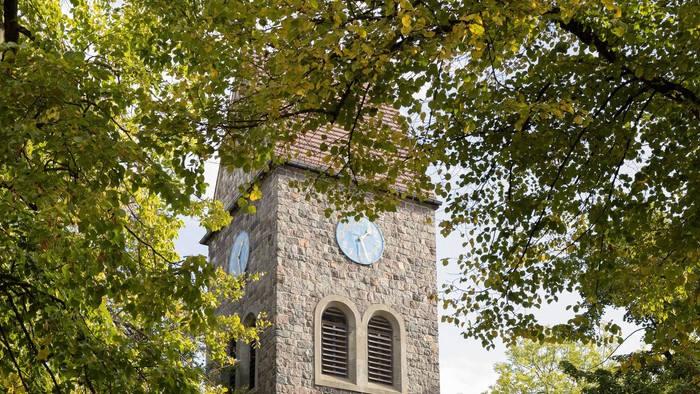 Der Kirchturm aus Stein mit Uhr und Fenster steht in einem Park.
