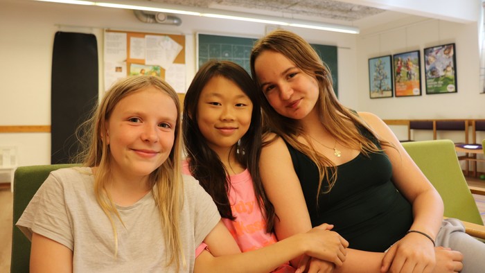 Tre muntre unge kvinder poser sammen i et klasseværelse eller studieområde.