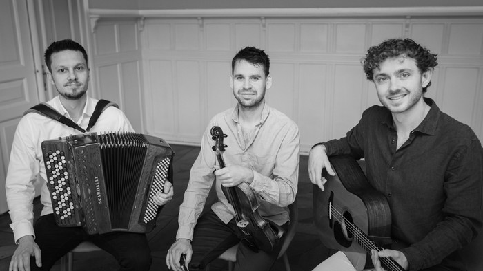 Tre musikere poser med harmonika, violin og guitar i et stilfuldt indendørs miljø.