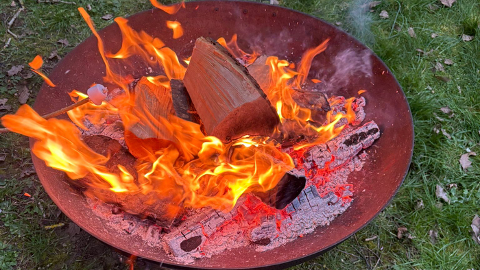 Ein tragbarer Grill mit glühender Kohle und Holzscheiten auf einer grasbewachsenen Fläche im Freien.