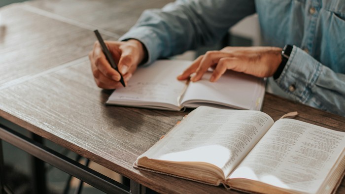 Person schreibt in Notizbuch neben aufgeschlagenem Buch
