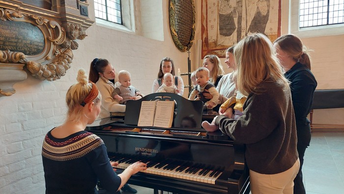 En kvinde spiller piano mens en gruppe af børn og voksne synger i en historisk sal.