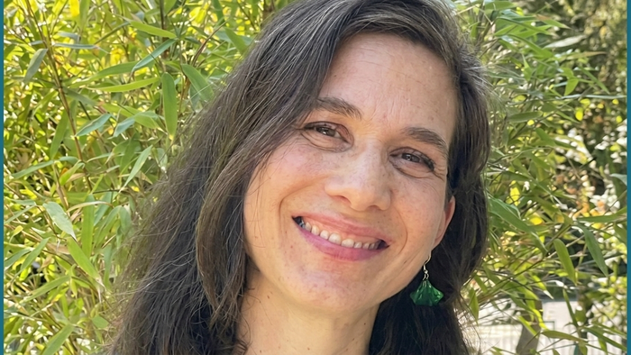 Ein Porträtfoto einer lächelnden Frau mit schulterlangen, braunen Haaren und grauen Strähnen. Sie trägt ein grünes Oberteil und einen grünen Ginkgoblatt-Ohrring. Der Hintergrund ist ein unscharfes, sonniges Grün von Bambuspflanzen. Das Foto ist links und 
