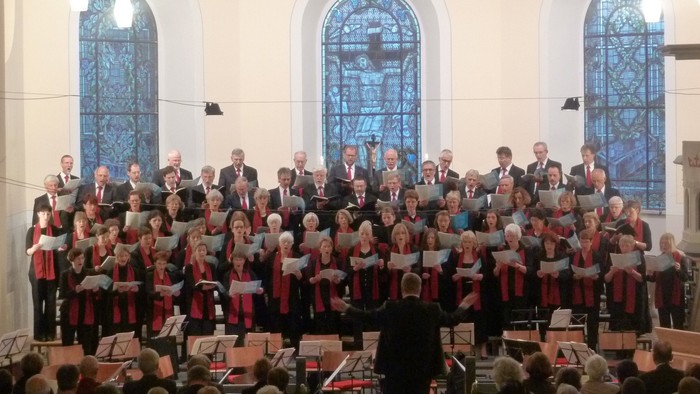 Ein großer Chor singt in einer Kirche, dirigiert von einem Mann im schwarzen Anzug
