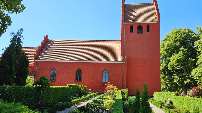 Livligt rødt kirkebyggeri med frodige haver og en klar blå himmel som baggrund.