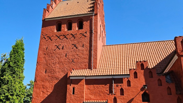 Livligt rød middelalderlig kirke med stejlt tag og dekorativt murværk under klar blå himmel.