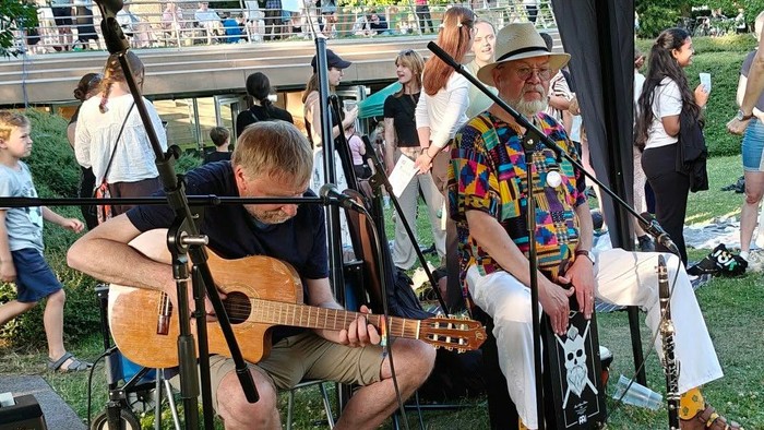 To musikere spiller guitar på scenen ved en udendørs begivenhed med et opmærksomt publikum.