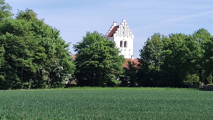 En fredfyldt landskabsscene med et højt kirketårn midt i frodige grønne marker og træer.