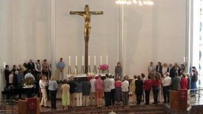 Ein modernes Kircheninneres mit Gläubigen, die sich um einen Altar versammeln, unter einem Kruzifix und bunten Kirchenfenstern.