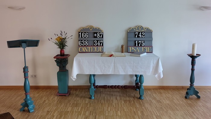 Minimalistischer Altarraum mit Kerzen, Tafeln und einfachen Holzstühlen in einem hellen Raum.