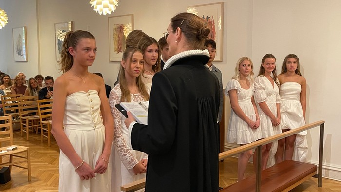 En gruppe unge piger i hvidt tøj står i en sal og lytter til en mand i mørkt tøj, der holder et papir.
