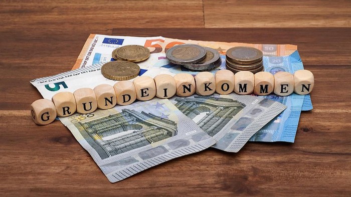 Holzklötze bilden das Wort „GrundEinkommen“, umgeben von Euro-Scheinen und Münzen, die auf einer Holzfläche angeordnet sind.