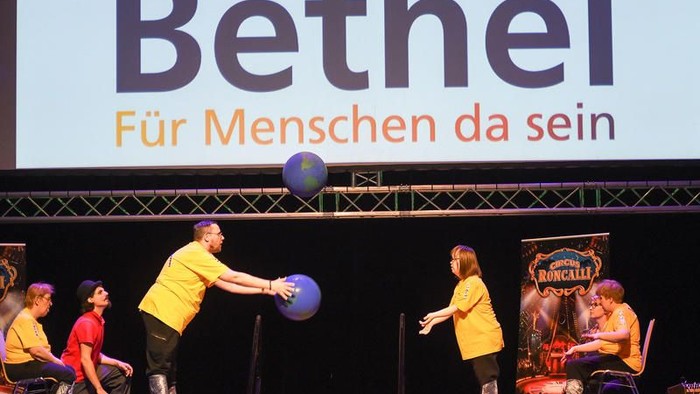 Eine Bühnenveranstaltung mit Darstellern, die mit einem großen blauen Ball unter dem Schild „Bethel – Für Menschen da sein“ spielen.