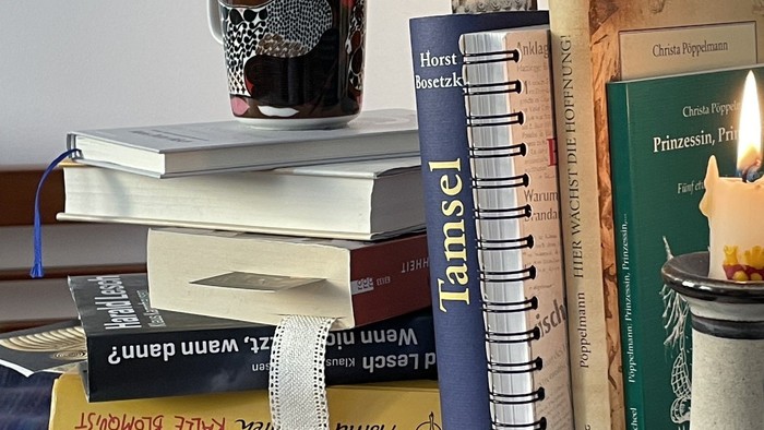Ein gemütlicher Stapel Bücher mit einer Tasse und einer Kerze auf einem Regal, der eine Leseecken-Atmosphäre vermittelt.