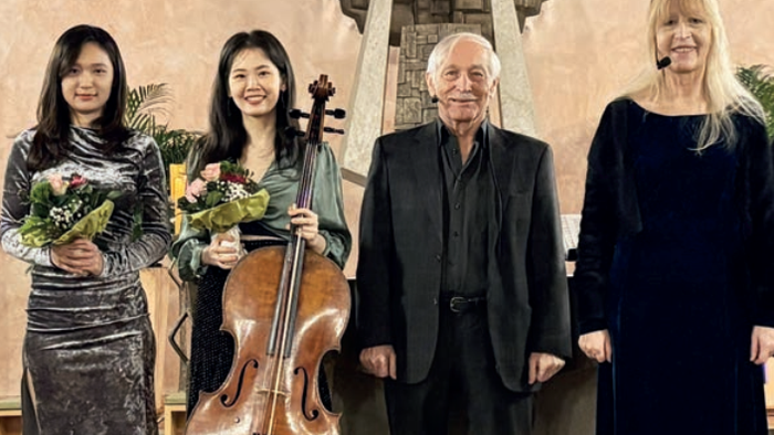 Vier Personen posieren gemeinsam, halten Blumen und ein Cello, wobei ein Mann in der Mitte steht und drei weitere ihn flankieren.
