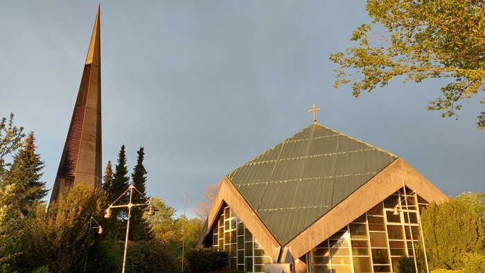 Moderne Kirche mit dreieckigem Dach und hohem, spitzem Turm in einer ruhigen Landschaft.