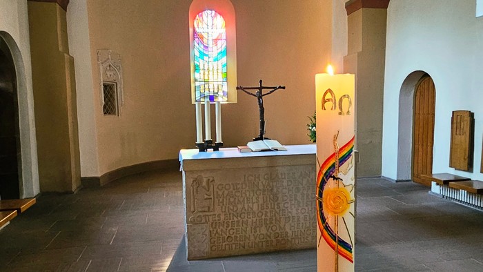 Ruhiger Kapelleninnenraum mit Buntglasfenstern, Altar, Kerze und schlichten Holzbänken.