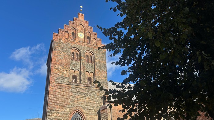 Historisk murstenskirketårn med buede vinduer og ur, indrammet af løvtræer under klar himmel.