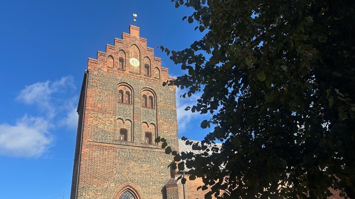 Historisk murstenskirketårn med buede vinduer og ur, indrammet af løvtræer under klar himmel.