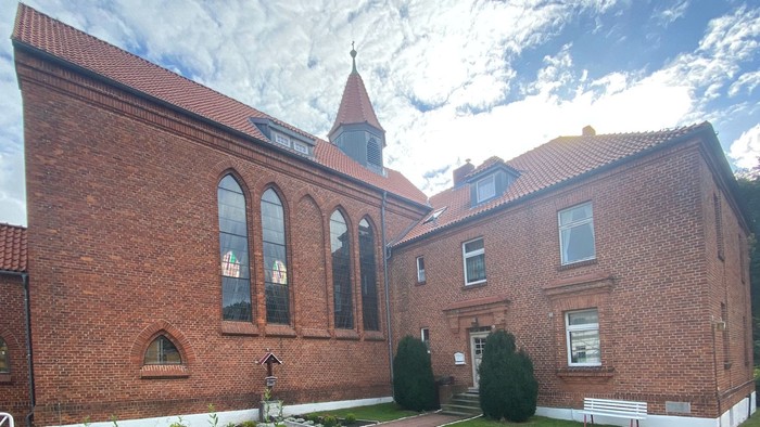 Ein rotes Backsteingebäude mit spitzem Turm und bogenförmigen Fenstern unter einem bewölkten Himmel.