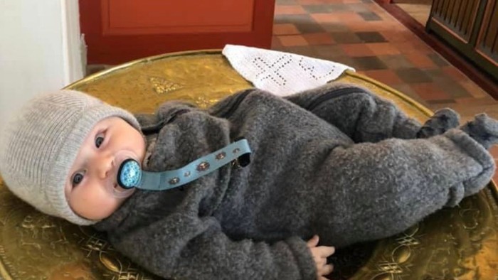 Et børnelev i graa klæder og hvid trøje ligger i en guld skål i kirke.