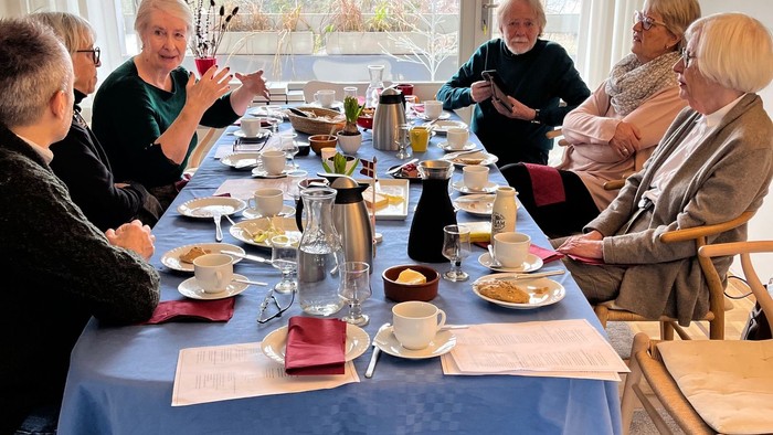 En gruppe mennesker, der nyder en social te-sammenkomst omkring et langt bord med forskellige snacks og drikkevarer.