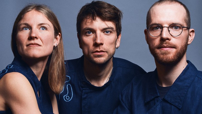 Tre personer i matchende marineblå outfits står tæt sammen mod en neutral baggrund.