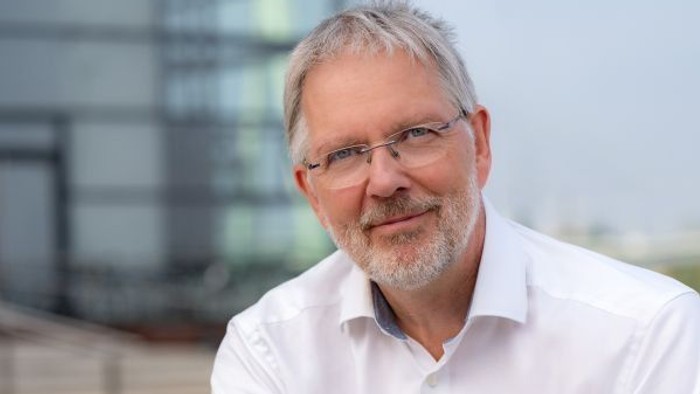 Ein lächelnder Mann mit Brille in einem weißen Hemd, der im Freien in der Nähe moderner Gebäude steht.