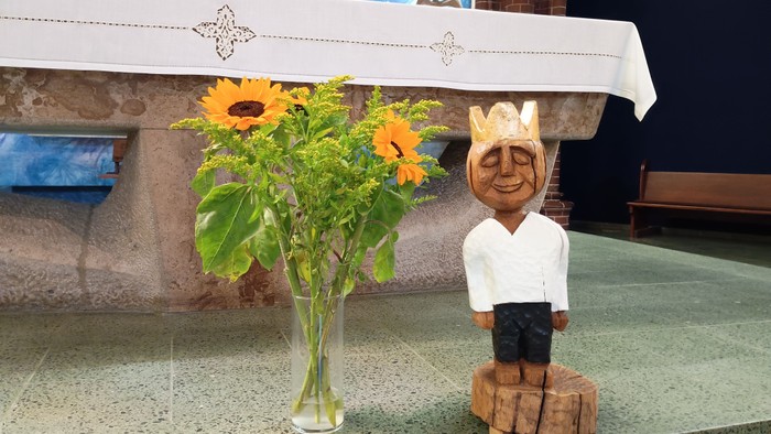 Eine hölzerne Statue einer lächelnden Figur neben einer Vase mit Sonnenblumen auf einem Kirchenaltar.