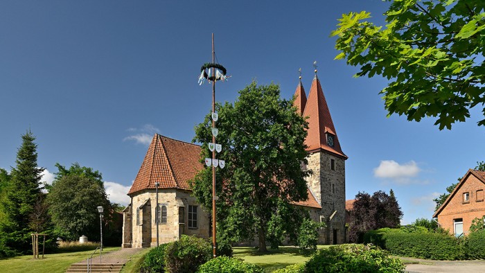 Historische Kirche mit hohem Kirchturm, umgeben von üppigem Grün und traditioneller Architektur unter klarem Himmel.