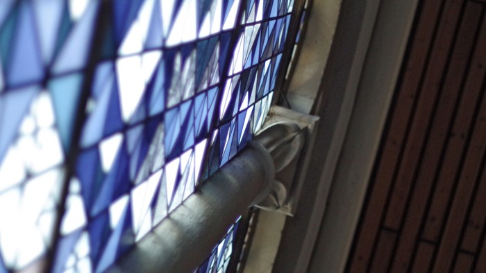 Eine Hand öffnet einen teilweise geschlossenen Holzladen und enthüllt den Blick auf buntes Kirchenfensterglas.