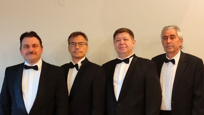 Vier Männer in formellen schwarzen Smokings posieren gemeinsam für ein professionelles Gruppenfoto.