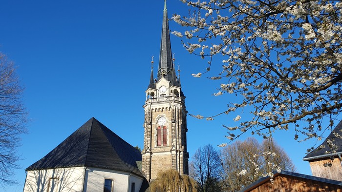 Eine friedvolle Kirche mit hohem Kirchturm steht neben blühenden Bäumen unter einem klaren blauen Himmel.