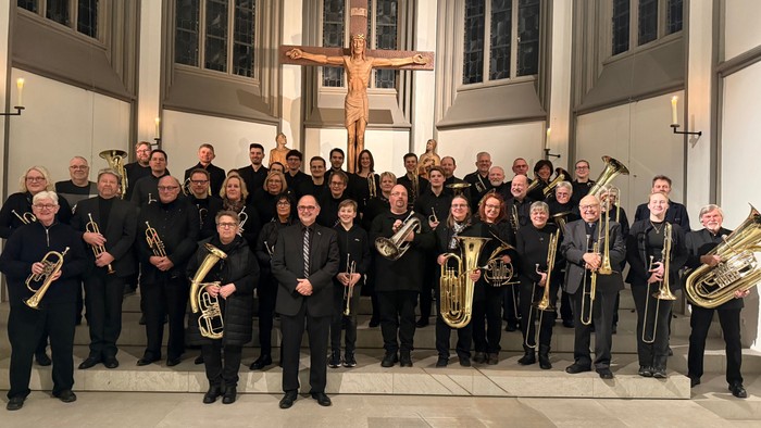 Eine formelle Orchestergruppe posiert gemeinsam in einer Kirche mit einem großen Kruzifix im Hintergrund.