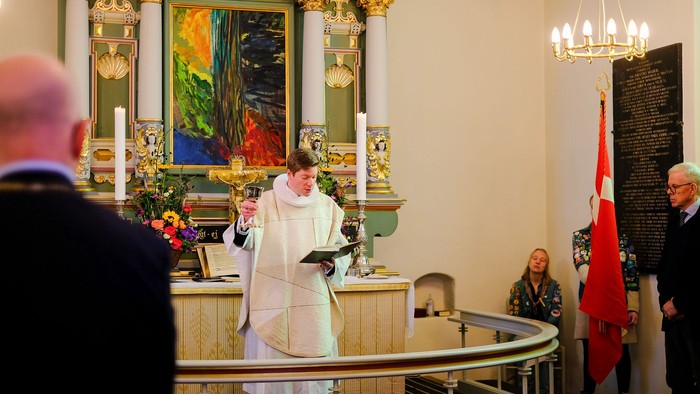 En præst leder en ceremoni i en kirke, hvor tilhørerne overværer det, og lysene er tændt.