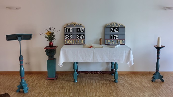 Minimalistischer Altarraum mit Kerzen, Tafeln und einfachen Holzstühlen in einem hellen Raum.