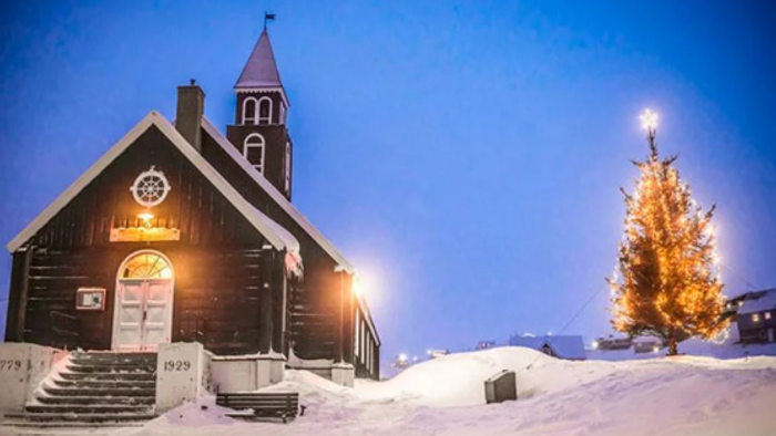 En snedækket trækirke med et tændt juletræ ved siden af, der gløder varmt mod vinternatten.