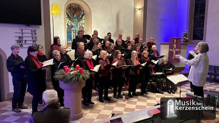 Der Chor singt unter der Leitung einer Dirigentin in einer festlich geschmückten Kirche vor einem Adventskranz mit vier roten Kerzen, während unten rechts das Logo der Konzertreihe „Musik bei Kerzenschein“ eingeblendet ist.