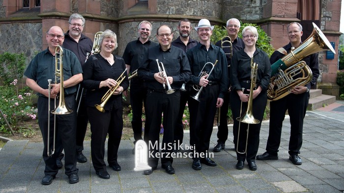 Das Blechbläserensemble posiert mit Trompeten, Posaunen, Tuba und weiteren Blechblasinstrumenten vor einer Kirche, während unten mittig das Logo der Konzertreihe „Musik bei Kerzenschein“ eingeblendet ist.