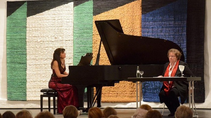 Zwei Frauen führen ein musikalisches Gespräch, zwischen ihnen steht ein Flügel in einem farbenfrohen, modernen Ambiente.