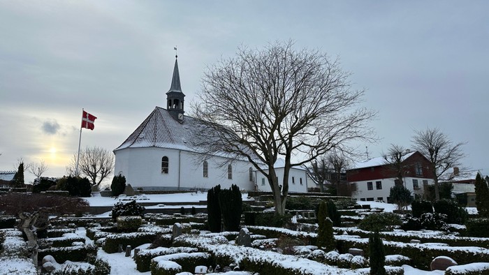 En fredfyldt vinterscene med en hvid kirke, formelle haver dækket af sne og et bladløst træ.