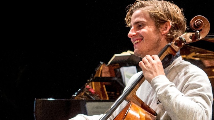 En musiker spiller cello med et glad udtryk, mens vedkommende sidder på scenen.