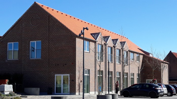 Moderne murstensbygning med rødt tegltag, glasindgang og parkerede biler på en brolagt gårdsplads.